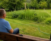 Mężczyzna siedzi na drewnianej ogrodowej huśtawce i patrzy na bujną łąkę kwietną wśród skoszonej trawy i drzew owocowych.