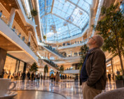 Mężczyzna stojący w wielopiętrowym atrium galerii handlowej zadziera głowę i spogląda ku wysokiemu szklanemu dachowi; na pierwszym planie widać stolik kawiarniany z filiżanką.